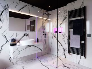 Une salle de bain moderne avec de grands carreaux blancs marbrés au mur et au sol, une douche à l’italienne en verre avec ciel de pluie, une niche éclairée d’une lumière colorée, un lavabo suspendu avec une vasque ronde et un chauffe-serviettes noir.